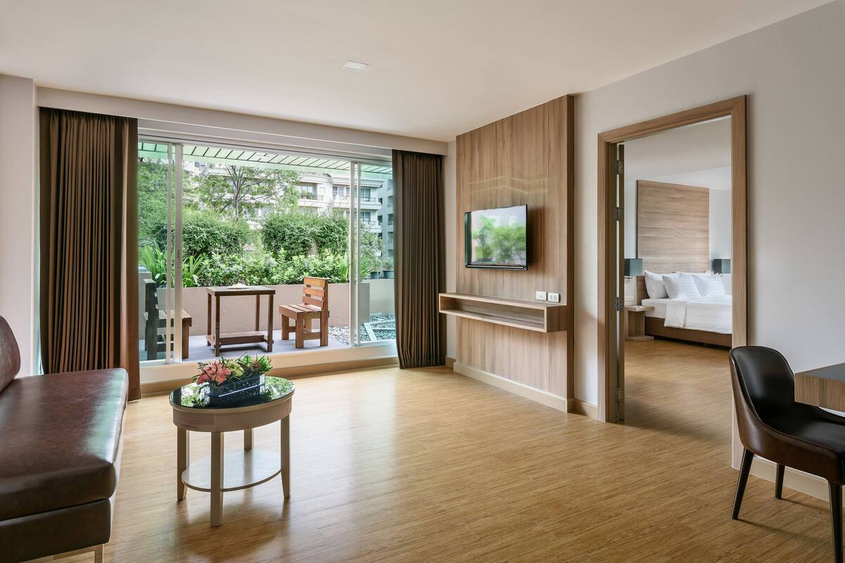 A modern hotel room with a wooden floor, a flat-screen TV mounted on the wall, a bed with white linens, and a balcony with a view of greenery.