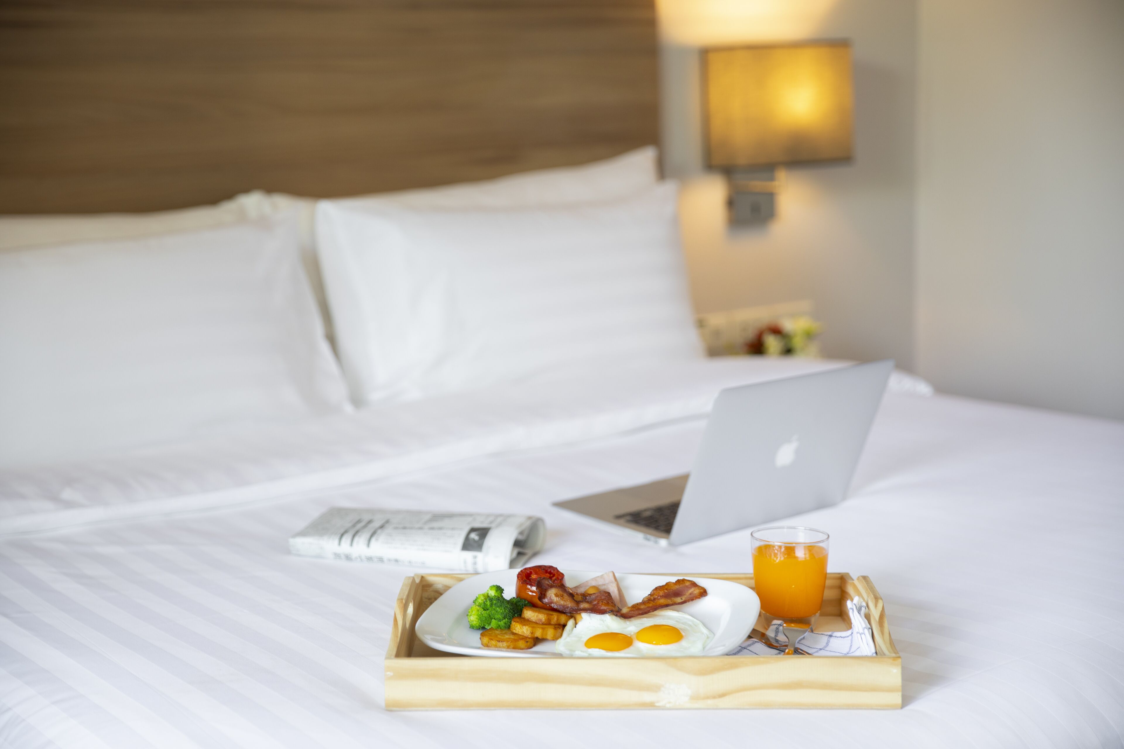 A neatly made bed with a laptop, a tray of breakfast including eggs, bacon, and toast, and a glass of orange juice.