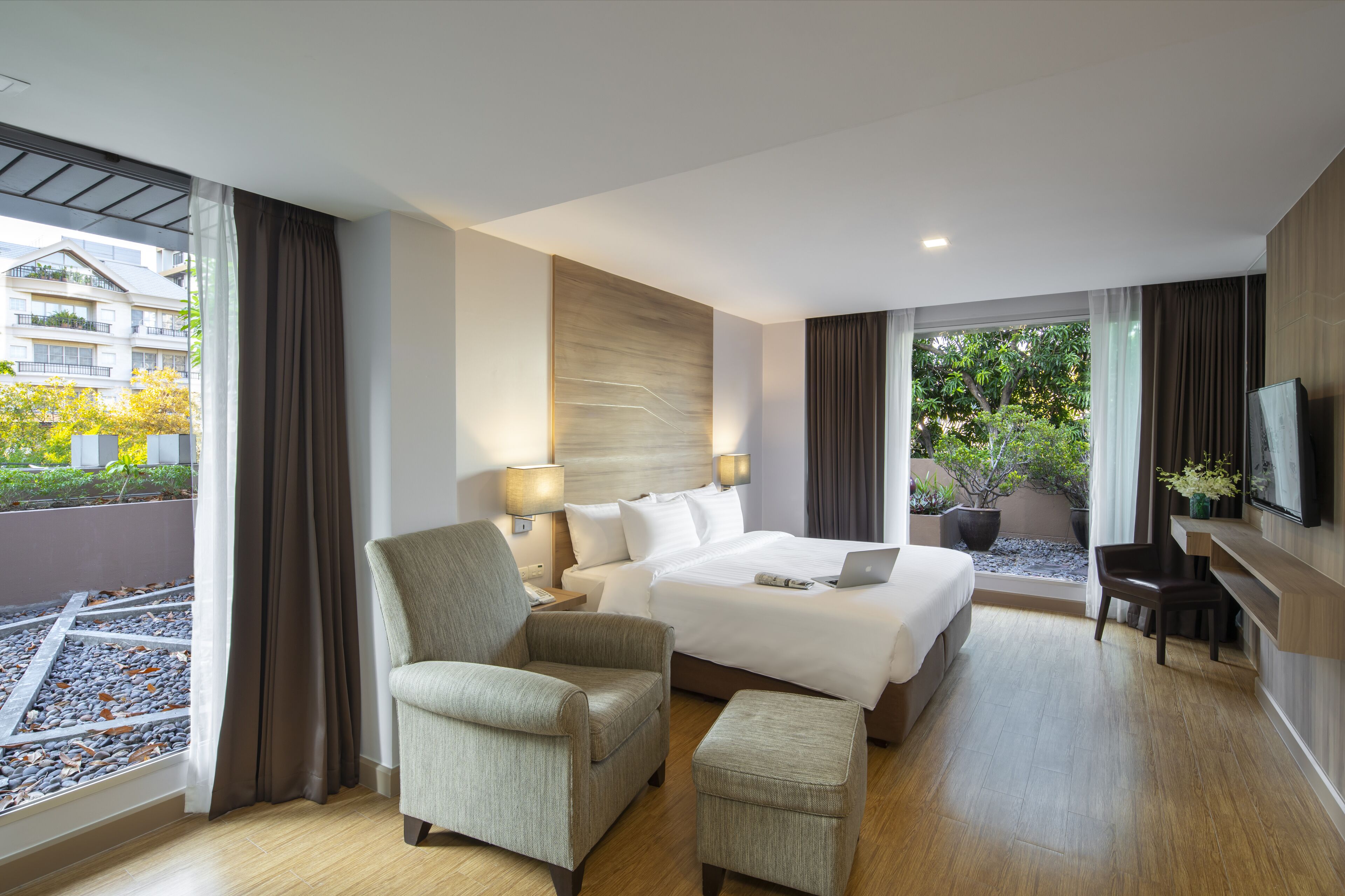 A hotel room with a large bed, a chair, a desk, and a TV. There are windows with curtains, a view of buildings outside, and a potted plant on the windowsill.