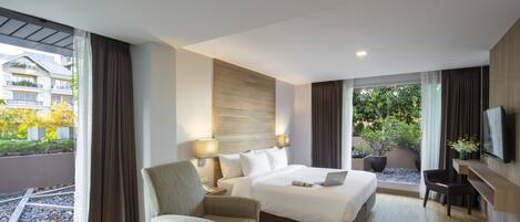Une chambre d’hôtel avec un grand lit, une chaise, un bureau et un téléviseur. Il y a des fenêtres avec des rideaux, une vue sur les bâtiments à l’extérieur et une plante en pot sur le rebord de la fenêtre.