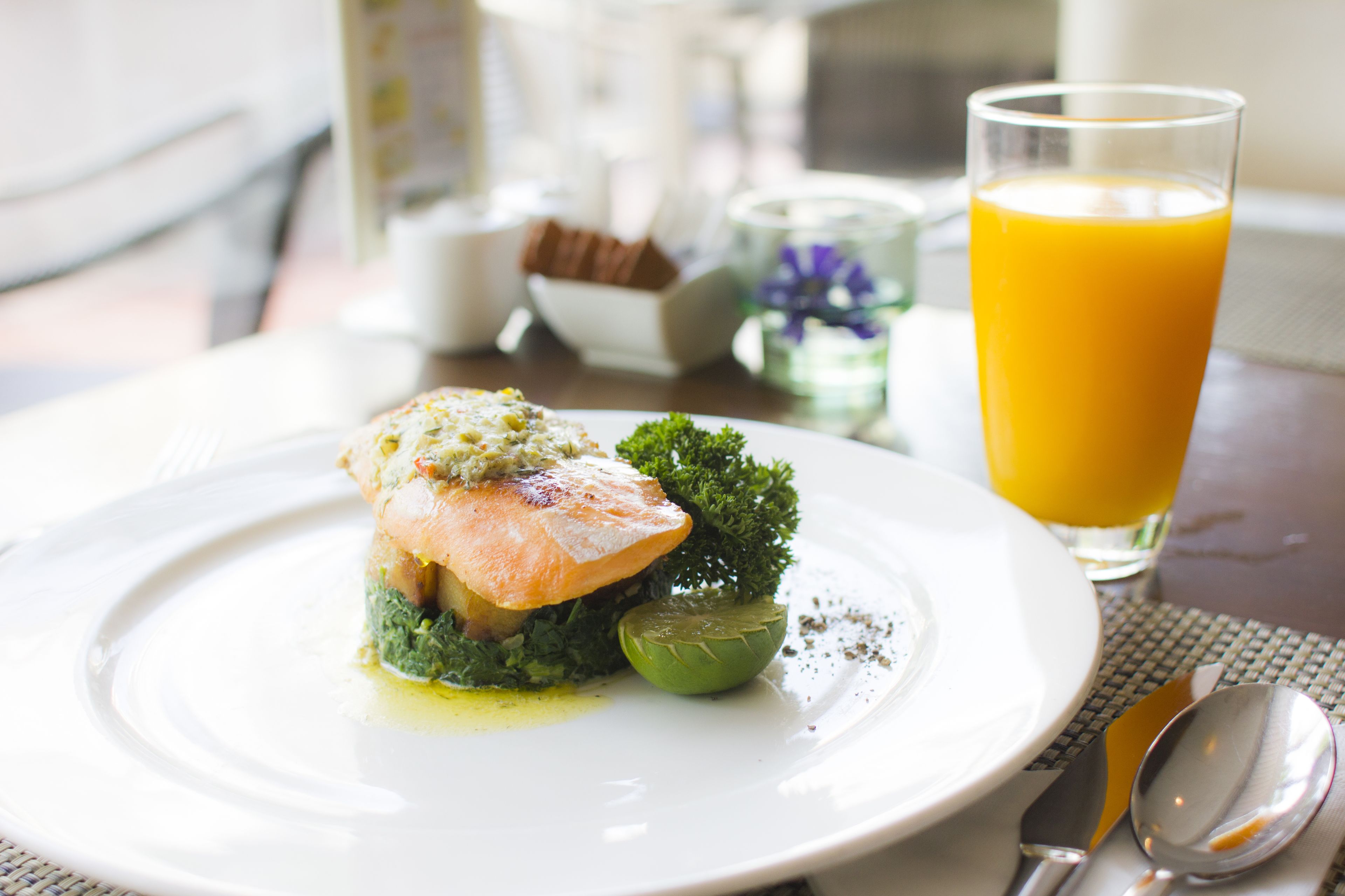 A plate of food with a piece of salmon and greens, a glass of orange juice, and a spoon and fork on a table.