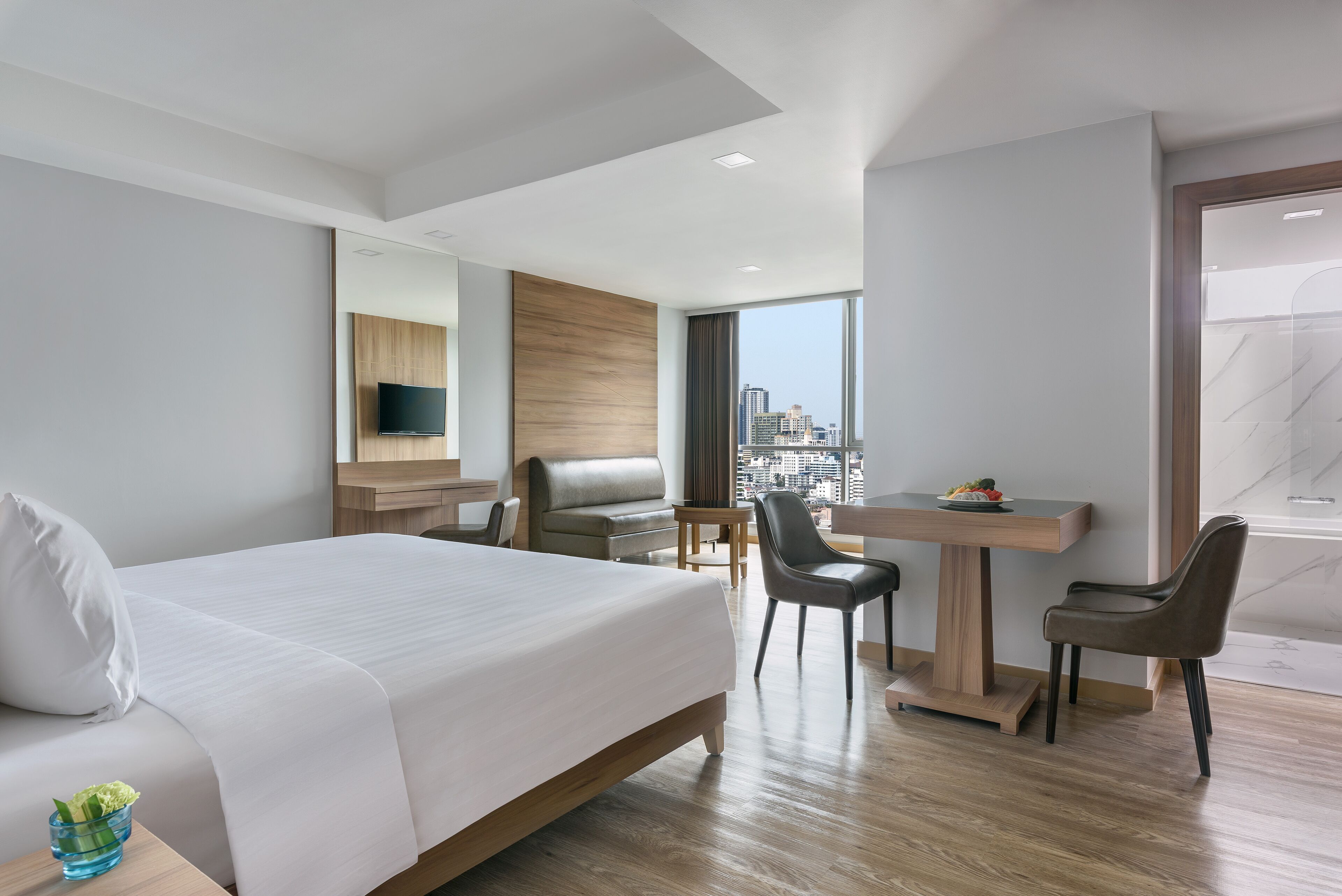 A modern hotel room with a large bed, a desk, two chairs, a TV, and a city view.