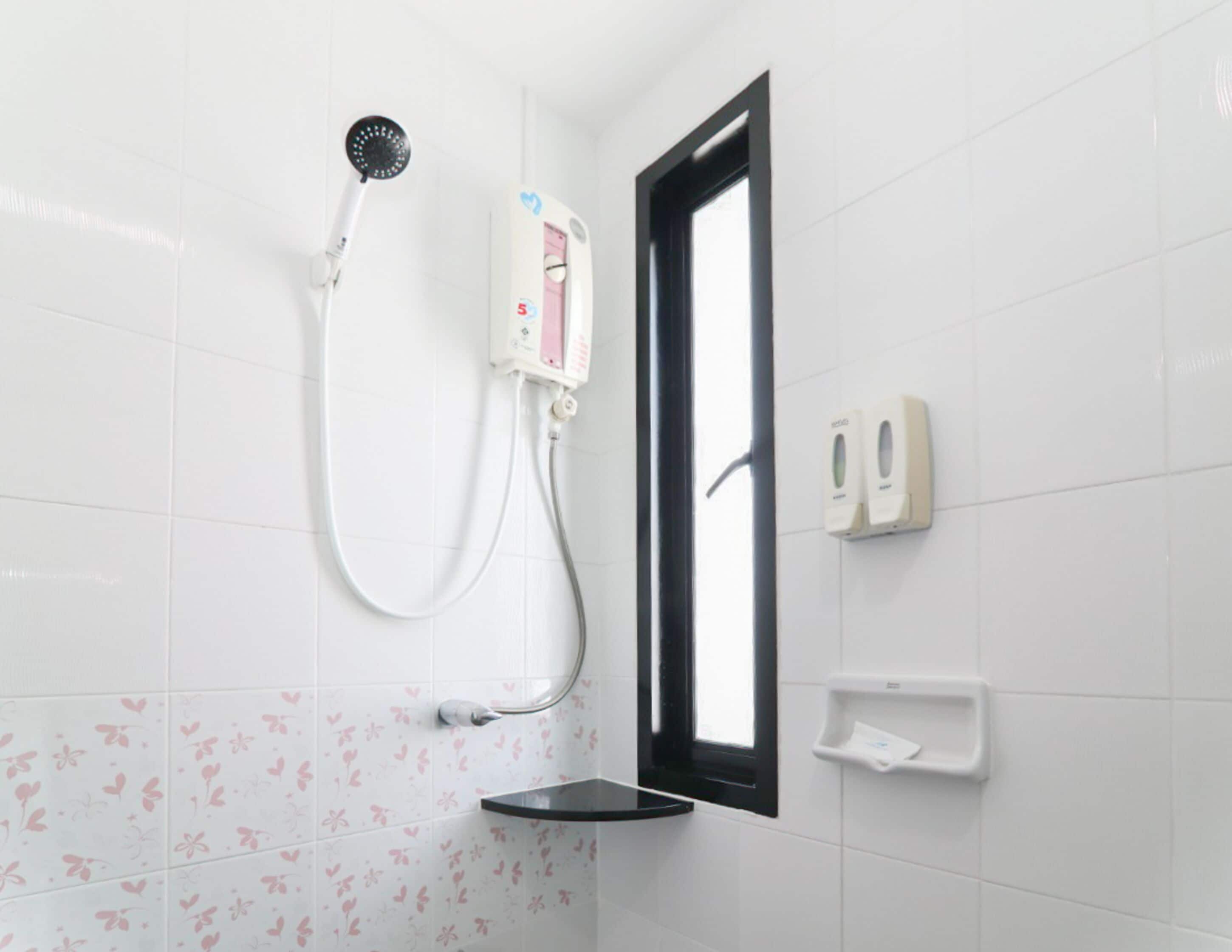 A bathroom with white tiles, a shower with a handheld showerhead, a window, and a soap dispenser.
