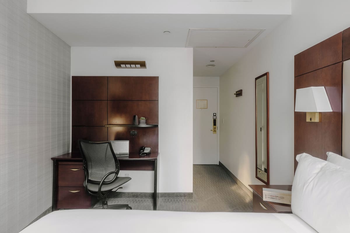 A hotel room with a bed, a desk with a chair, a wardrobe, a bedside table with a lamp, and a door.