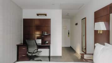 Habitación de hotel con cama, escritorio y silla, armario, mesita de noche con lámpara y una puerta.
