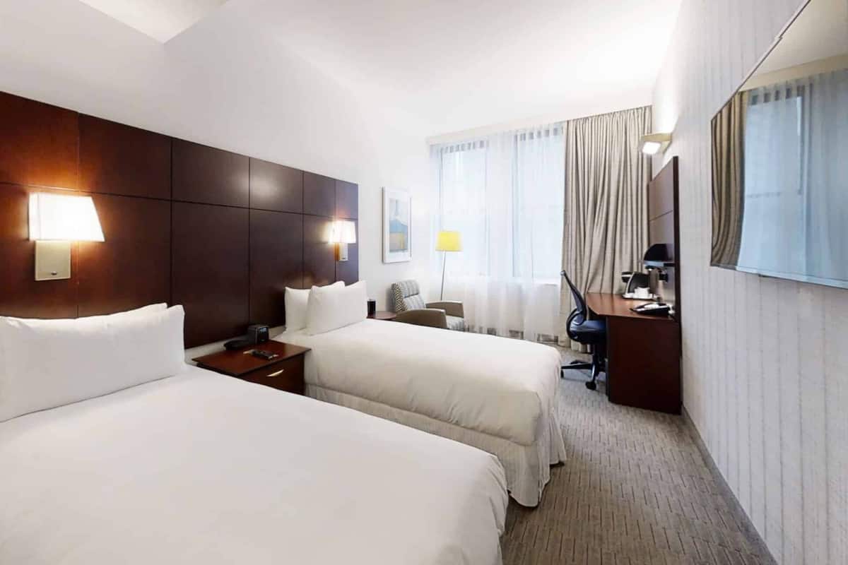 A hotel room with two beds, a desk, a chair, and a large window with curtains.