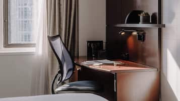 A hotel room with a bed, a desk with a chair, a coffee machine, and a wall-mounted shelf with a plant.