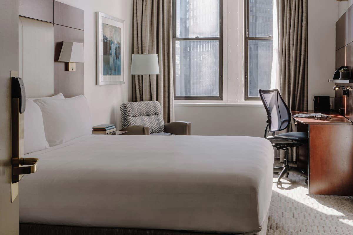 A hotel room with a large bed, a desk, a chair, a lamp, and a window with curtains.