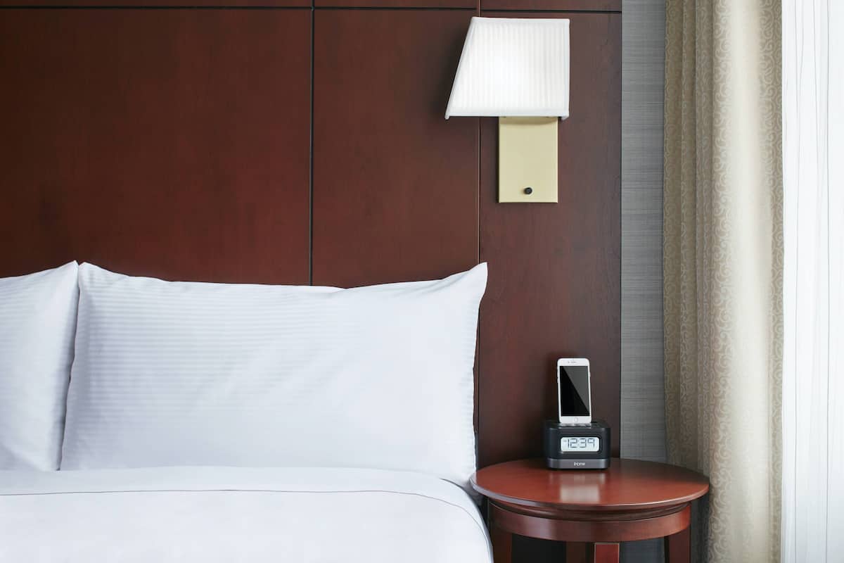 A hotel room with a bed, a bedside table with a clock, a wall-mounted lamp, and a curtain.
