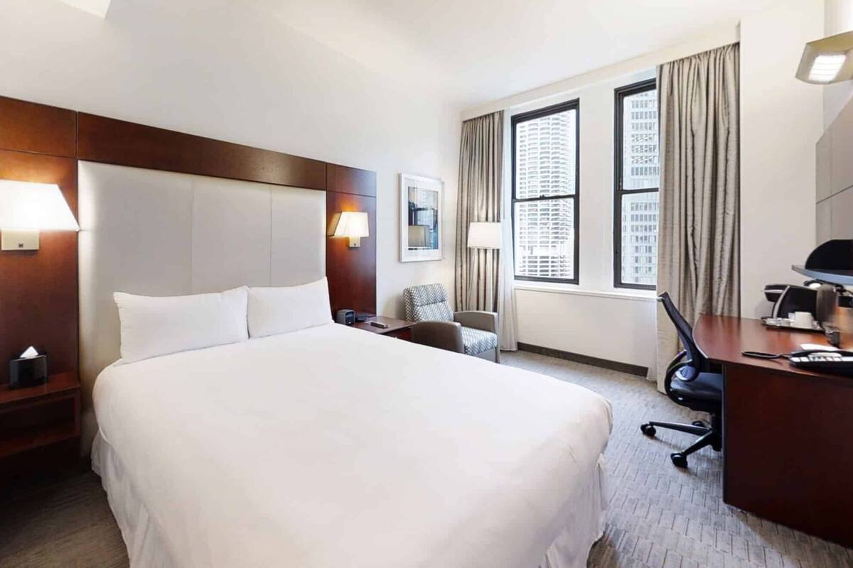 A hotel room with a large bed, a desk, a chair, a lamp, and a window with curtains.