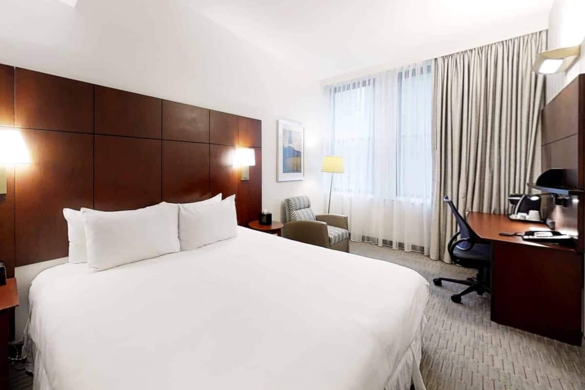 A hotel room with a large bed, a desk with a chair, a lamp, and a window with curtains.