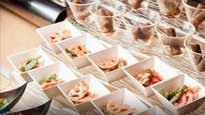 A table with multiple square white dishes containing food, and several clear glasses with a brown liquid, possibly tea.