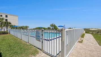 En inhägnad pool med kristallklart blått vatten, en vit byggnad och en asfalterad gångväg.