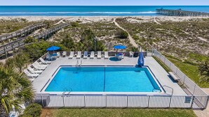 A rectangular pool with a wooden deck, surrounded by lounge chairs and umbrellas, with a beach and pier in the background. - Beachside Motel (Fernandina Beach)