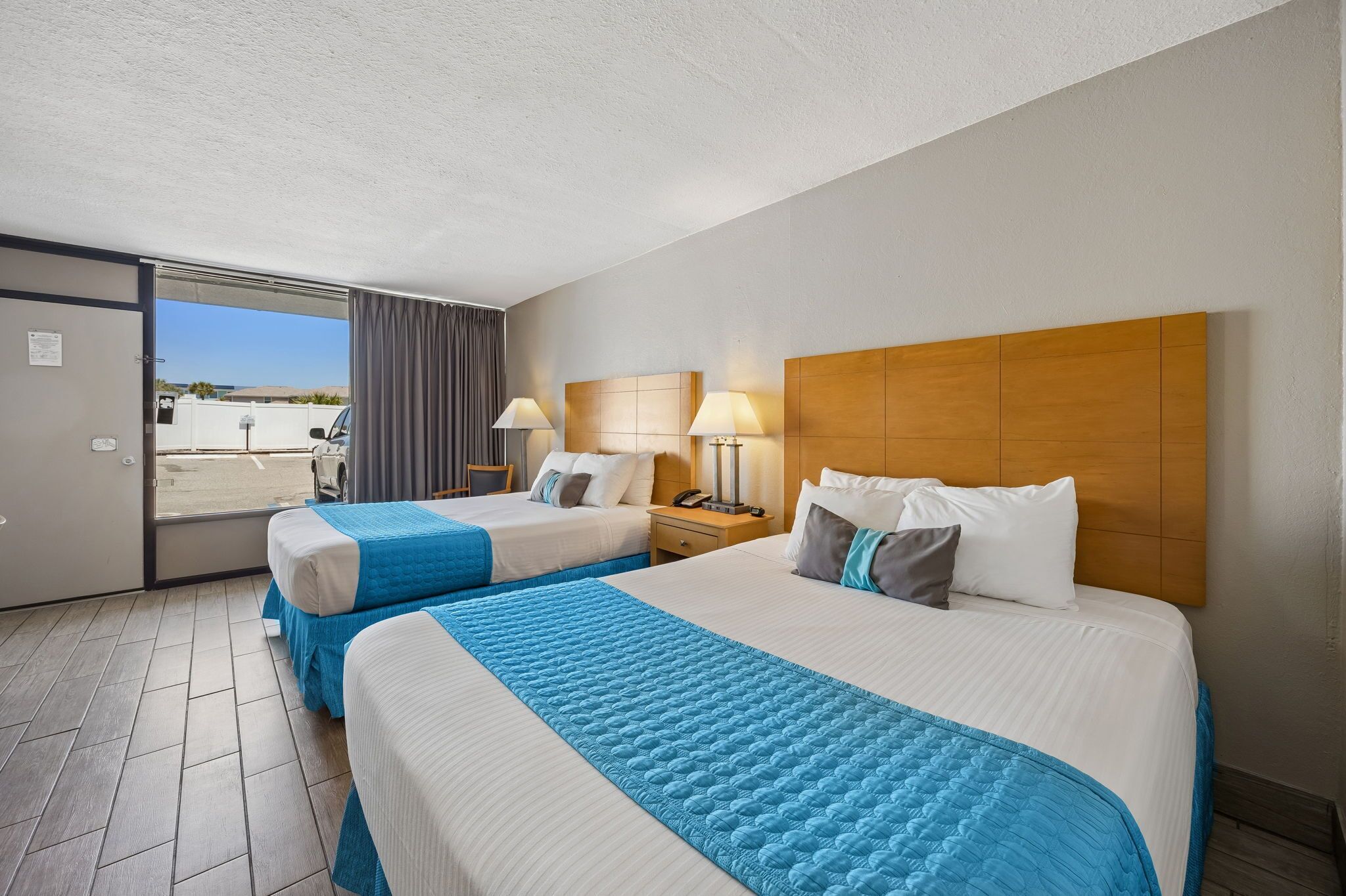 A hotel room with two beds, a wooden headboard, a nightstand with a lamp, and a view through a sliding door.