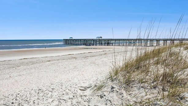 A beach with a pier extending into the water, sandy beach, and sparse vegetation. - Beachside Motel (Fernandina Beach)
