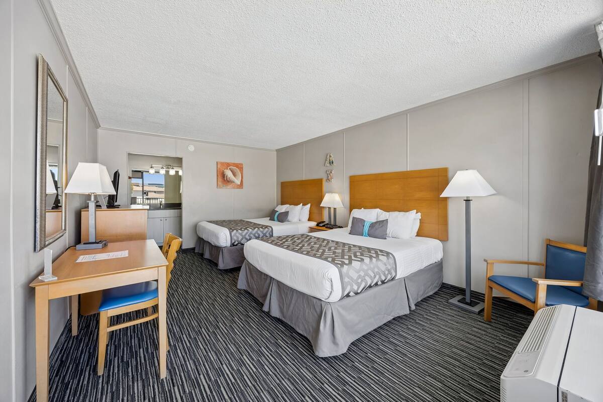 A hotel room with two beds, a desk, chairs, a mirror, and a lamp.