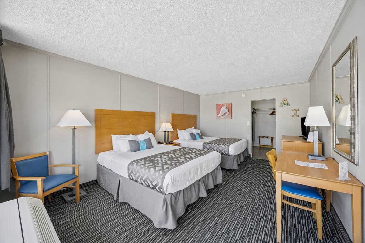 A hotel room with two beds, a desk, a chair, a lamp, and a mirror.