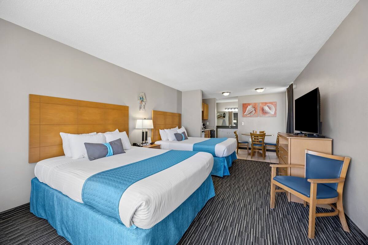 A hotel room with two beds, a desk, a chair, a television, and a kitchenette.