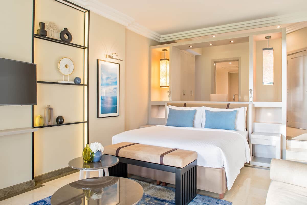 A hotel room with a large bed, bedside tables, a TV, a wall-mounted shelf with decorative items, and a view of the hallway.