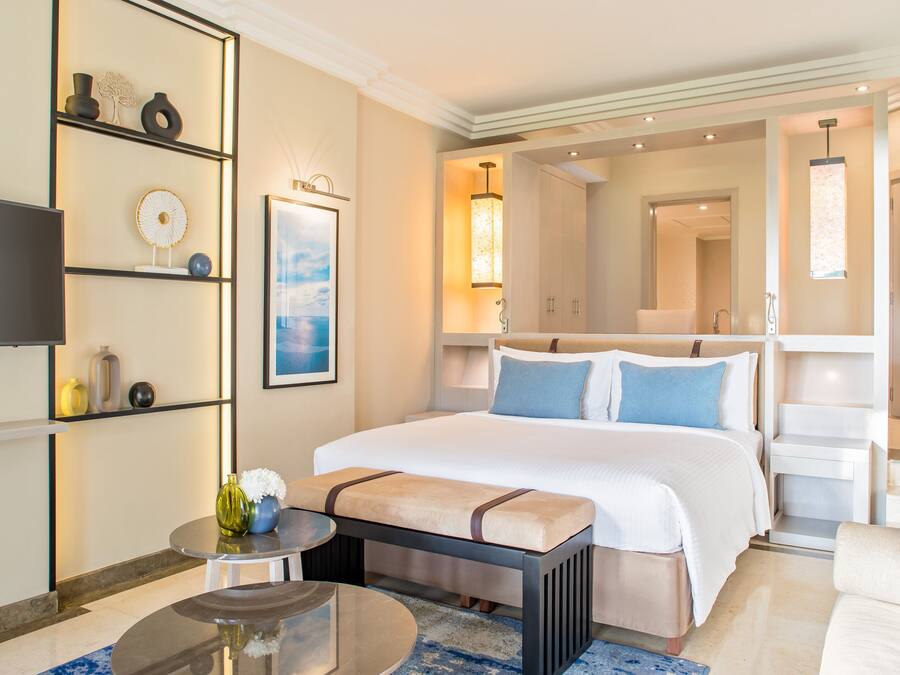 A hotel room with a large bed, bedside tables, a TV, a wall-mounted shelf with decorative items, and a view of the hallway.
