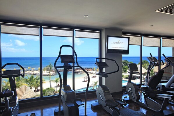 A gym with exercise equipment and a large window overlooking a beach and ocean.