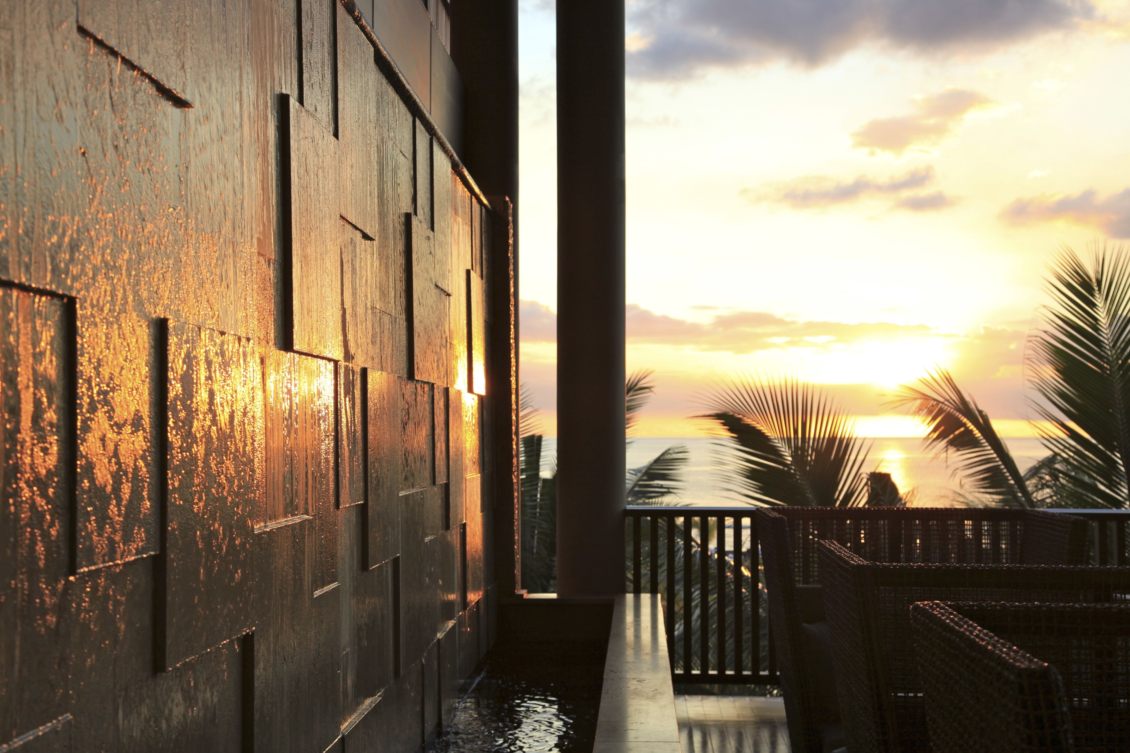 A wooden exterior wall with a water feature and a balcony with a view of the sunset and palm trees.