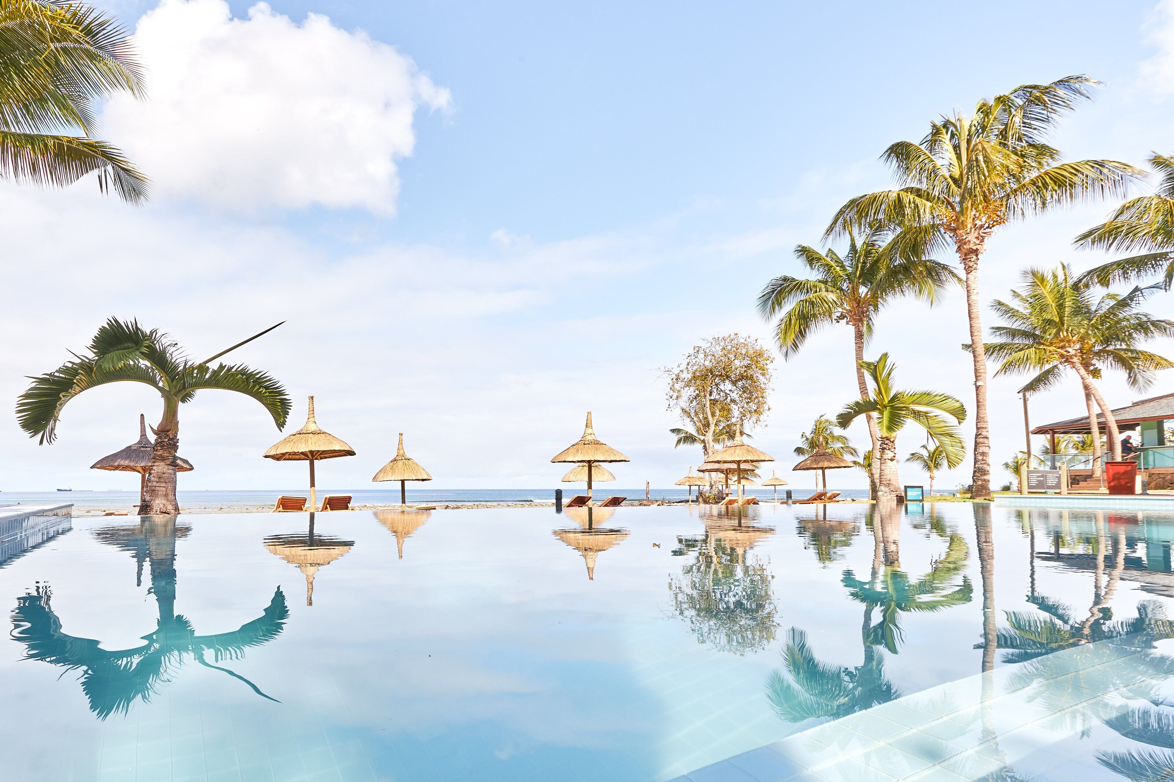 A clear view of a swimming pool with reflections of palm trees and umbrellas, surrounded by beach chairs and a building with a poolside bar.