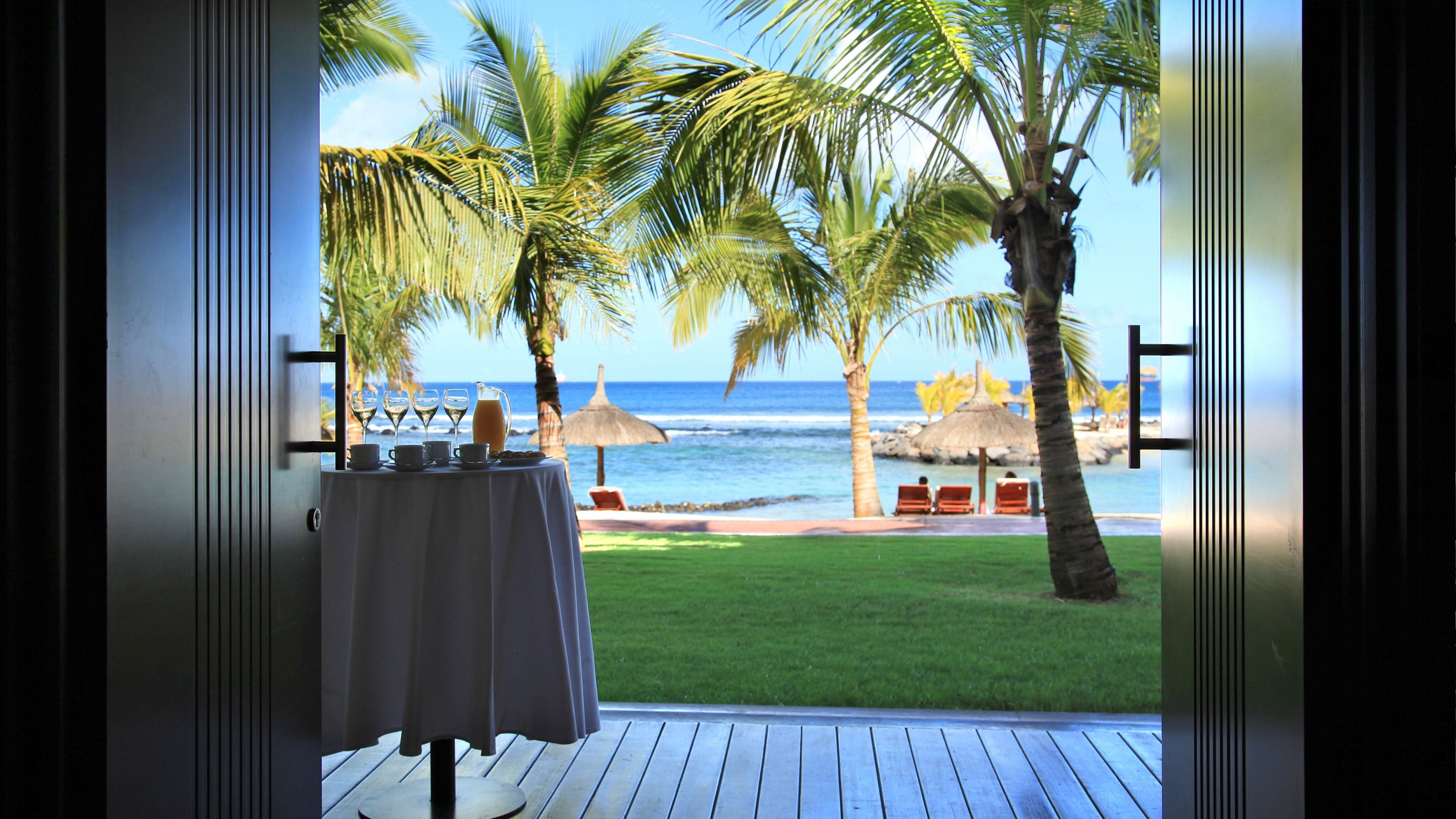 A view from a room with a table set for breakfast overlooking a beach with palm trees and the ocean.