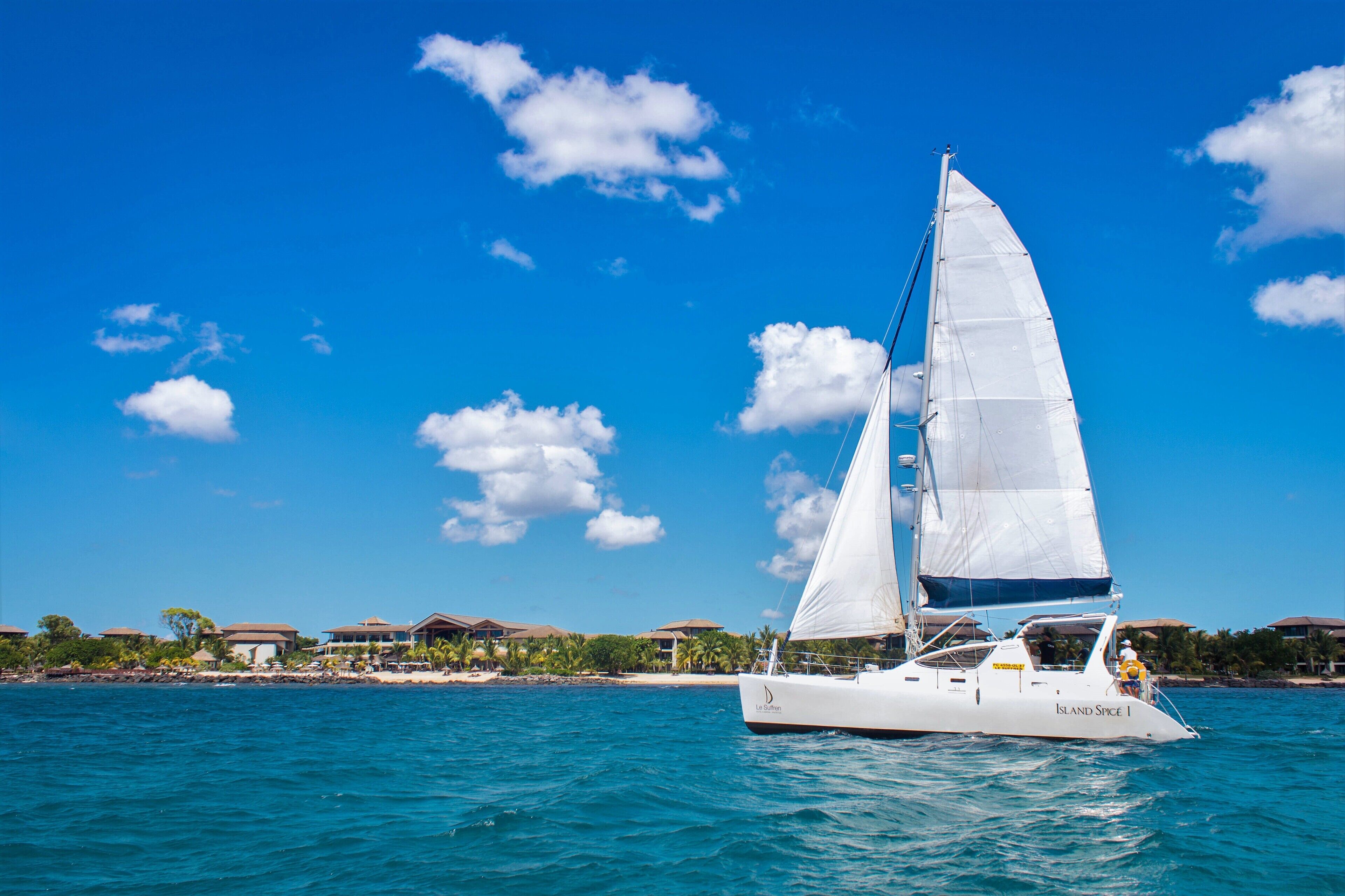 Een zeilboot genaamd "Island Spice 1" op helderblauw water, met op de achtergrond strandhuizen en palmbomen.