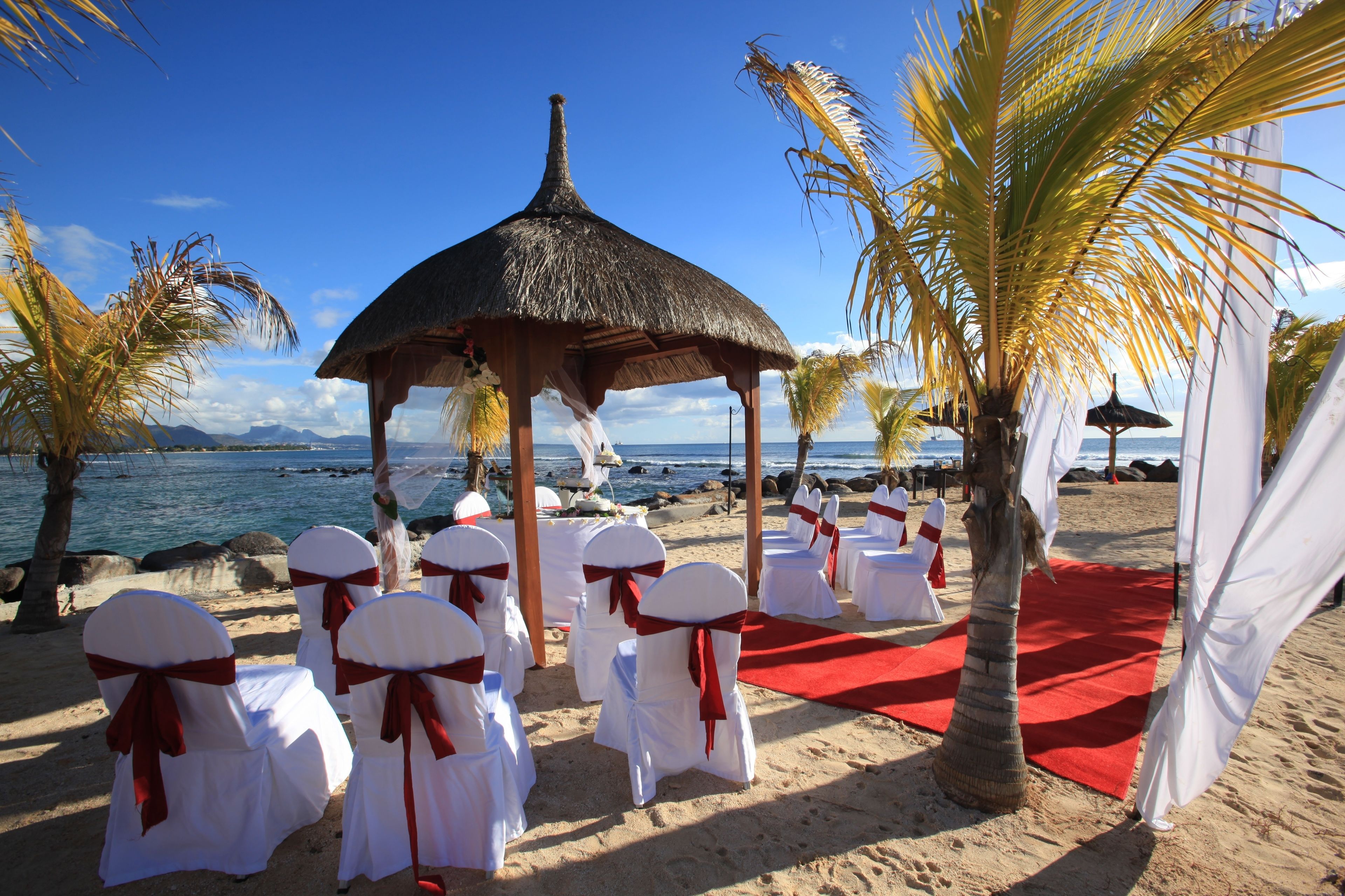 Montaje para una boda en la playa con sillas blancas y un pabellón de techo de paja.