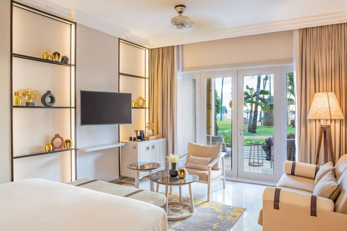 A modern hotel room with a bed, a TV, a seating area with a sofa and chairs, a small table, and a balcony with a view of palm trees.