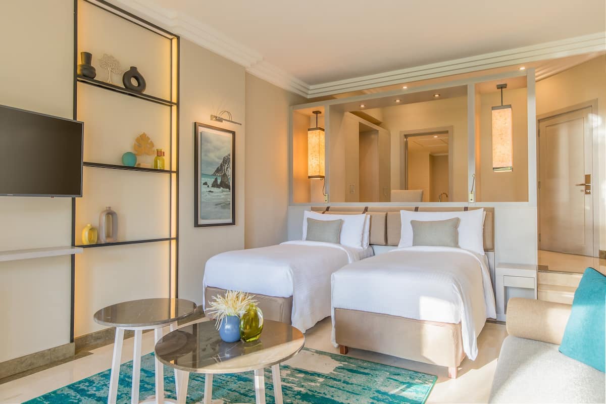 A hotel room with two beds, a TV, a small table with a vase, and a large mirror.