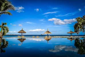 A serene infinity pool with four thatched umbrellas and chairs, reflecting the clear blue sky and calm sea.