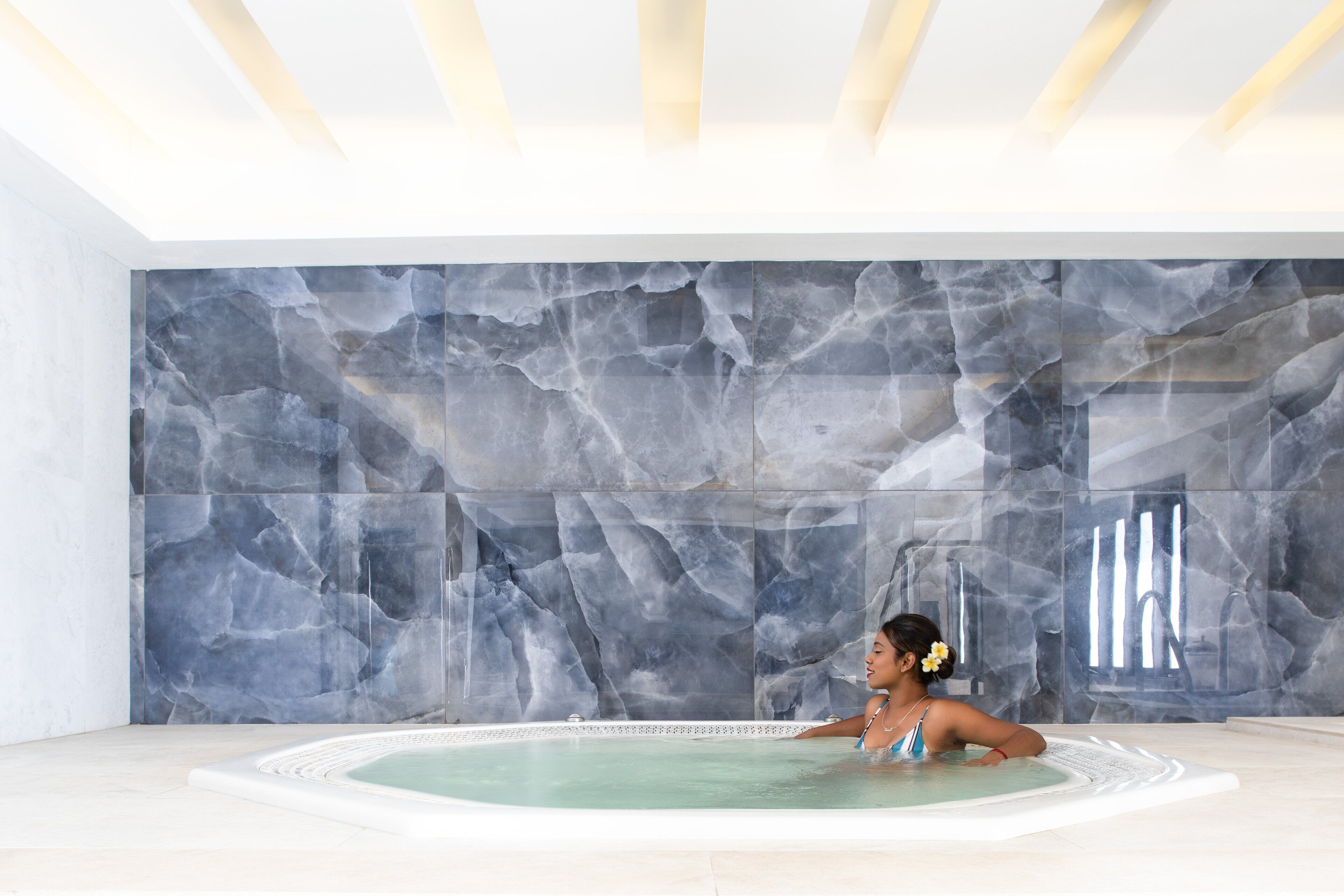 A woman relaxing in a hot tub with a marble wall backdrop.