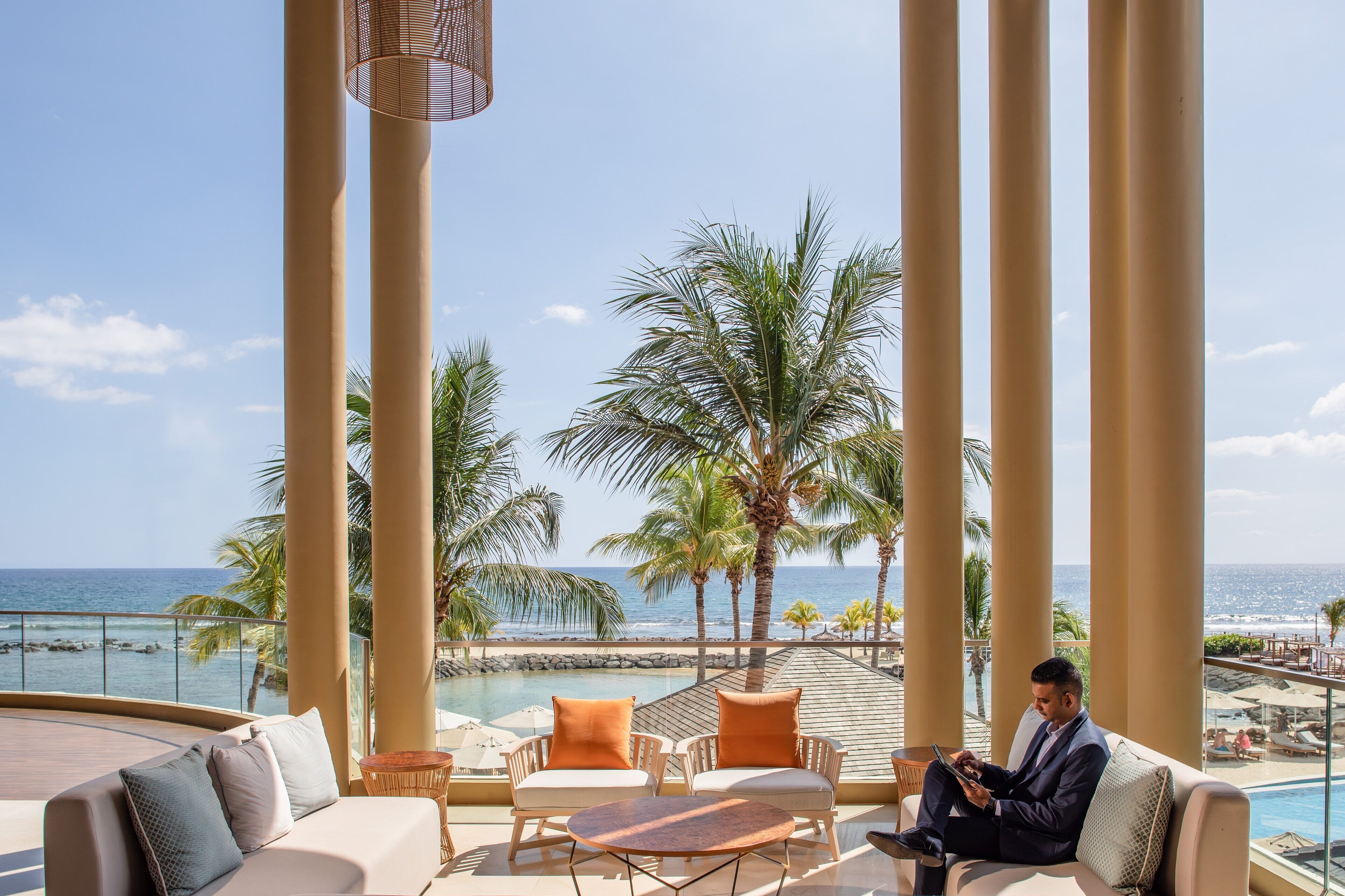 Un área de estar exterior lujosa con vista al océano, palmeras y una piscina.