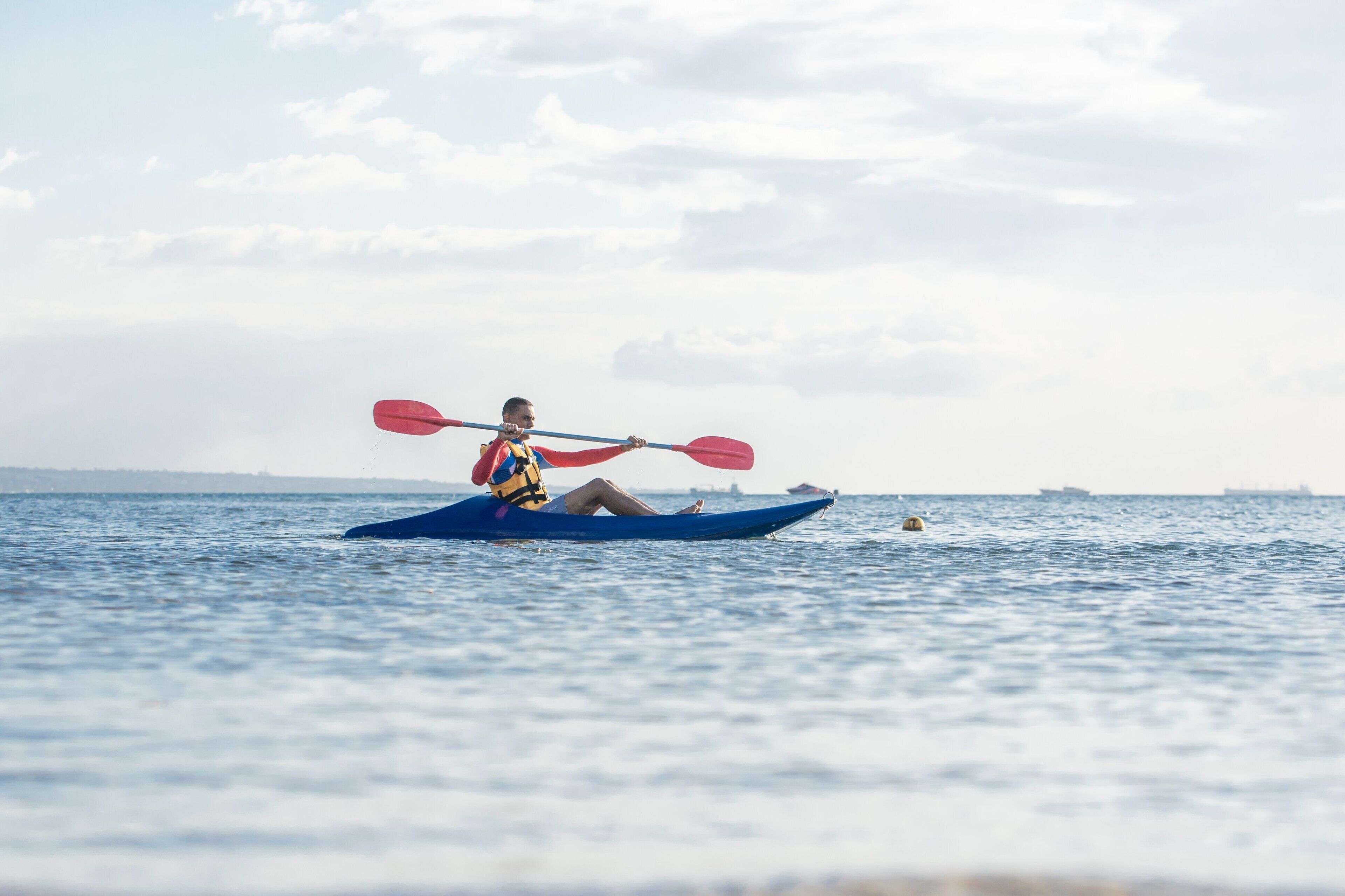 Una persona en kayak sobre aguas tranquilas, con una costa lejana y barcos al fondo.