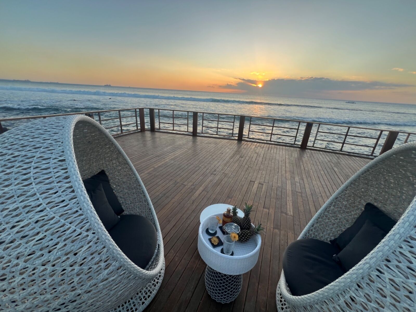 Un ponte di legno con sedie in vimini e un tavolino con bevande e snack, con vista sull'oceano al tramonto.