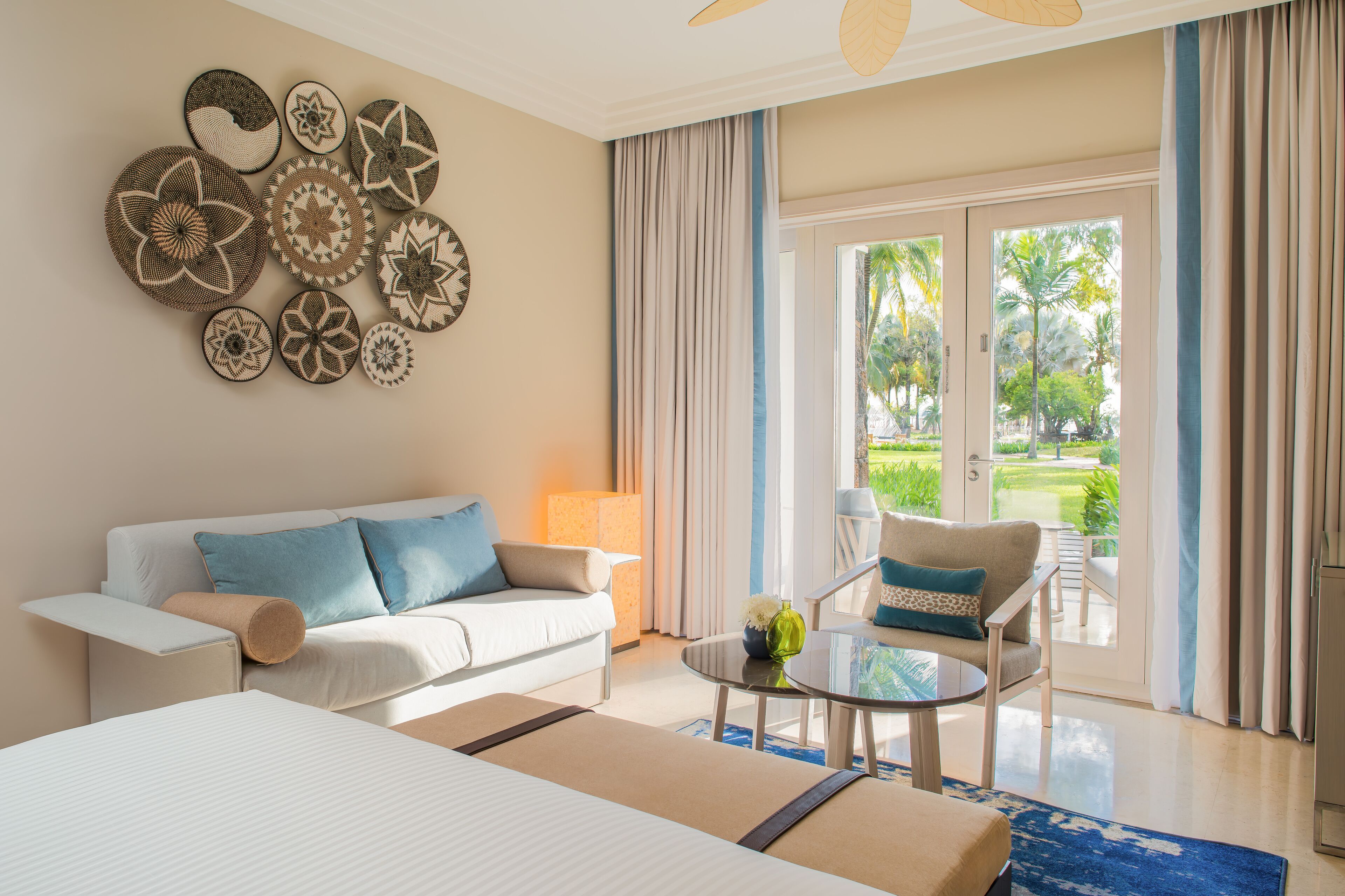 A hotel room with a bed, a sofa, a chair, a table with a vase, and a view of a garden through the door.