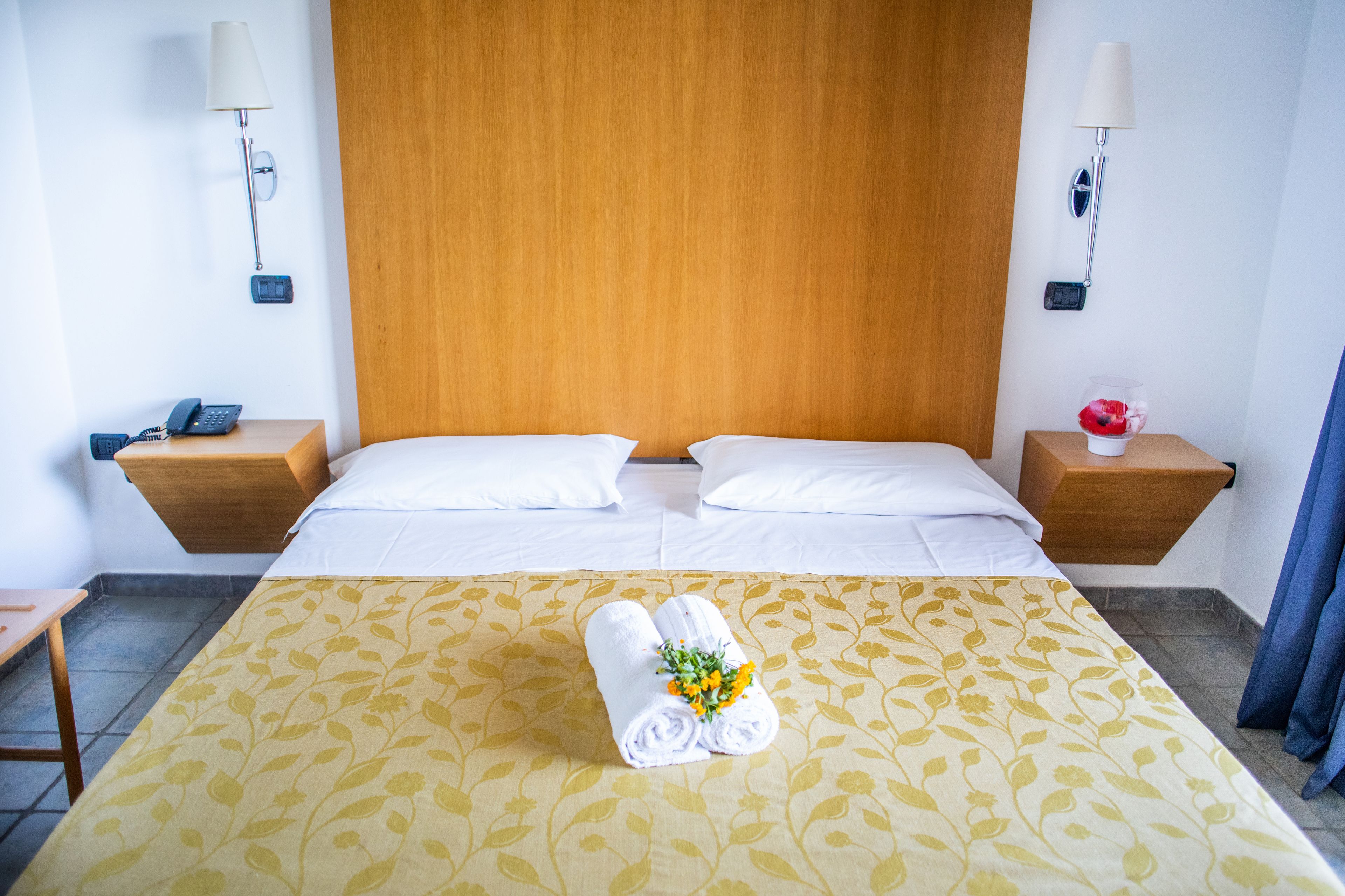 Une chambre d’hôtel avec une tête de lit en bois, deux lits avec du linge de lit blanc, une couvre-lit à motifs floraux, une table de chevet avec une lampe et une petite table avec un téléphone et un objet décoratif.