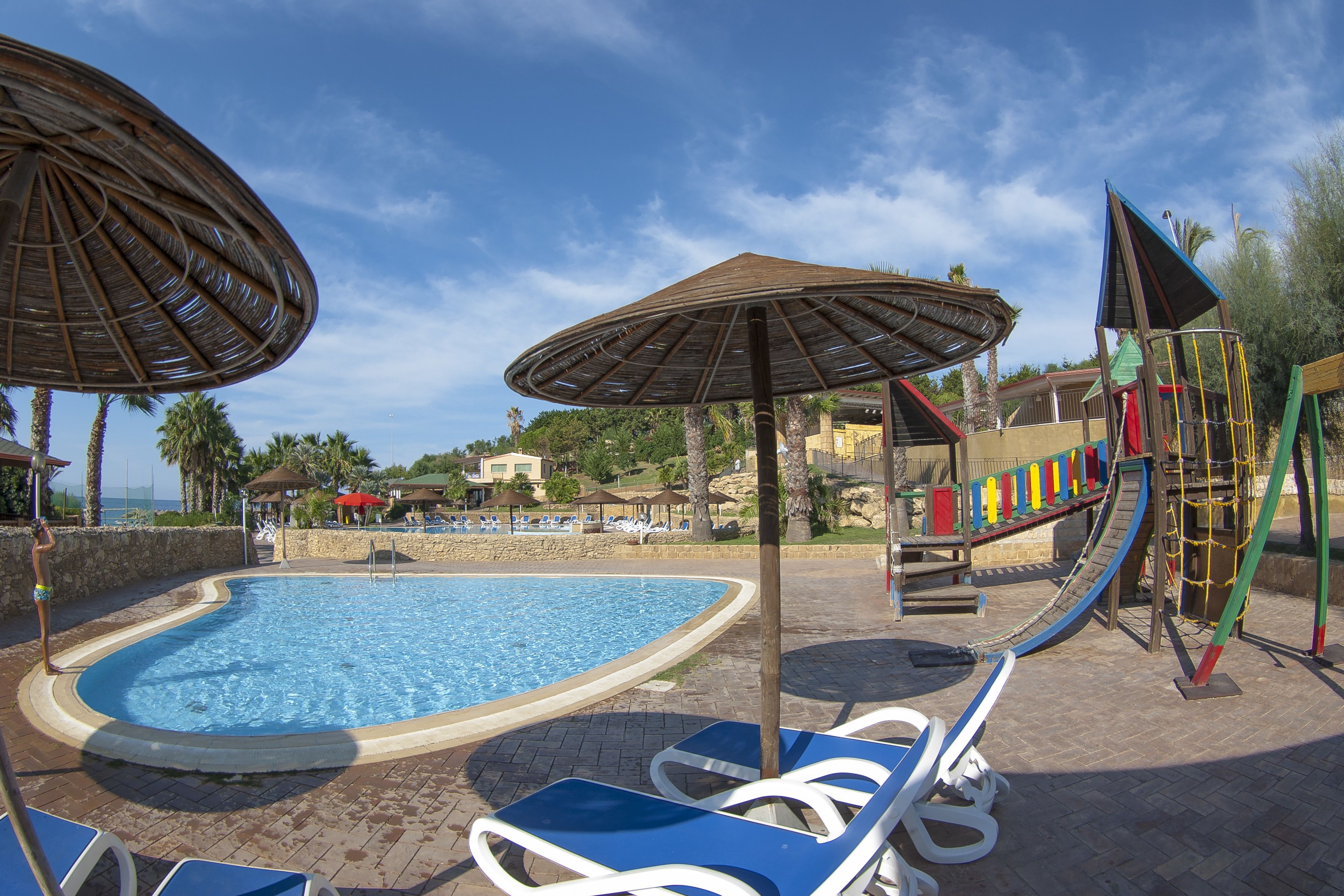 Un espace piscine avec des chaises longues et des parasols, une aire de jeux pour enfants et une vue sur le paysage environnant.