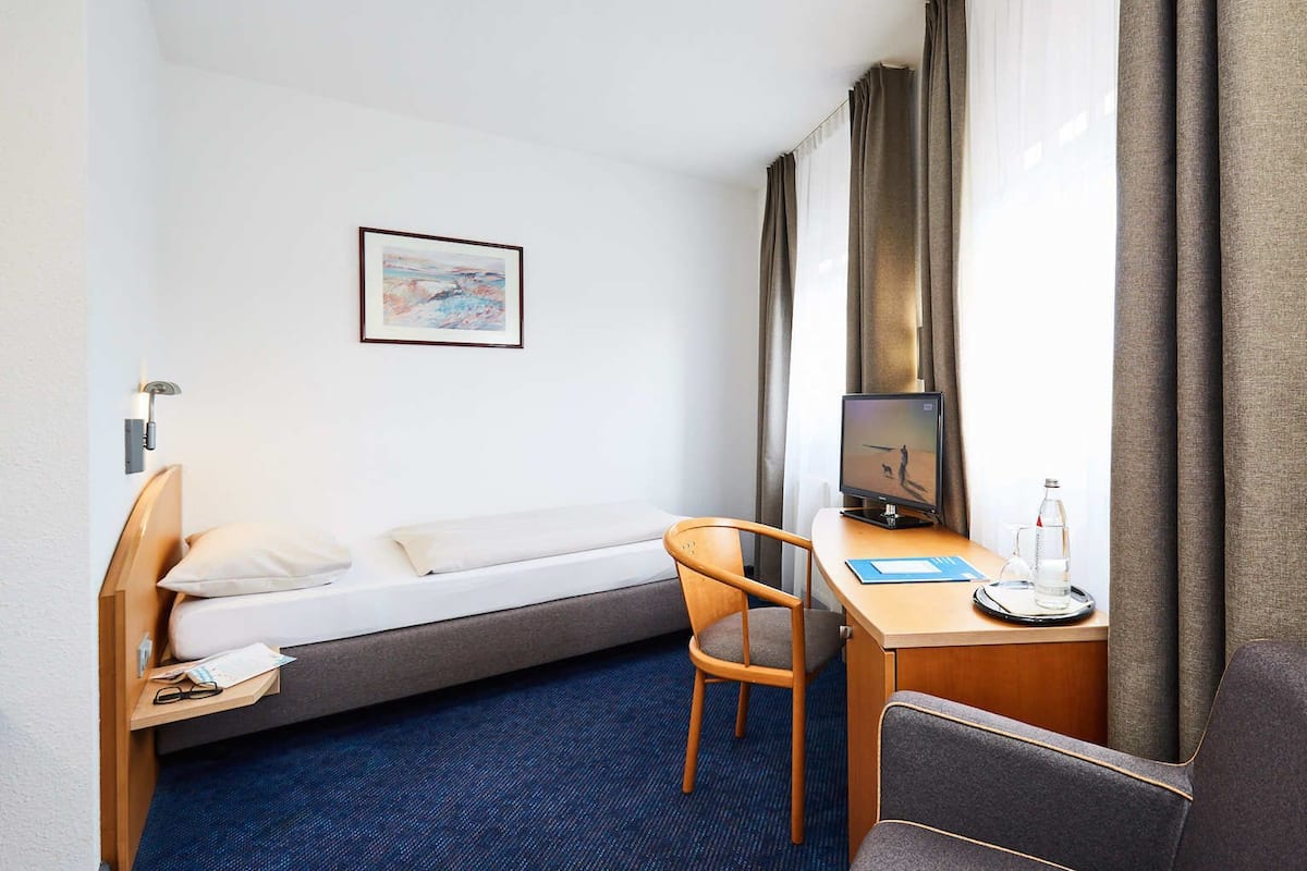 Hotellihuone, jossa on sänky, työpöytä tietokoneella, tuoli, pieni pöytä ja ikkuna, jossa on verhot.
