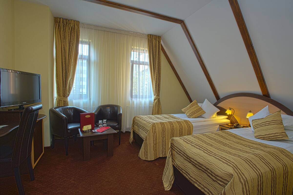 Habitación de hotel con dos camas, un escritorio con televisor, una silla y una mesa con una carpeta roja.