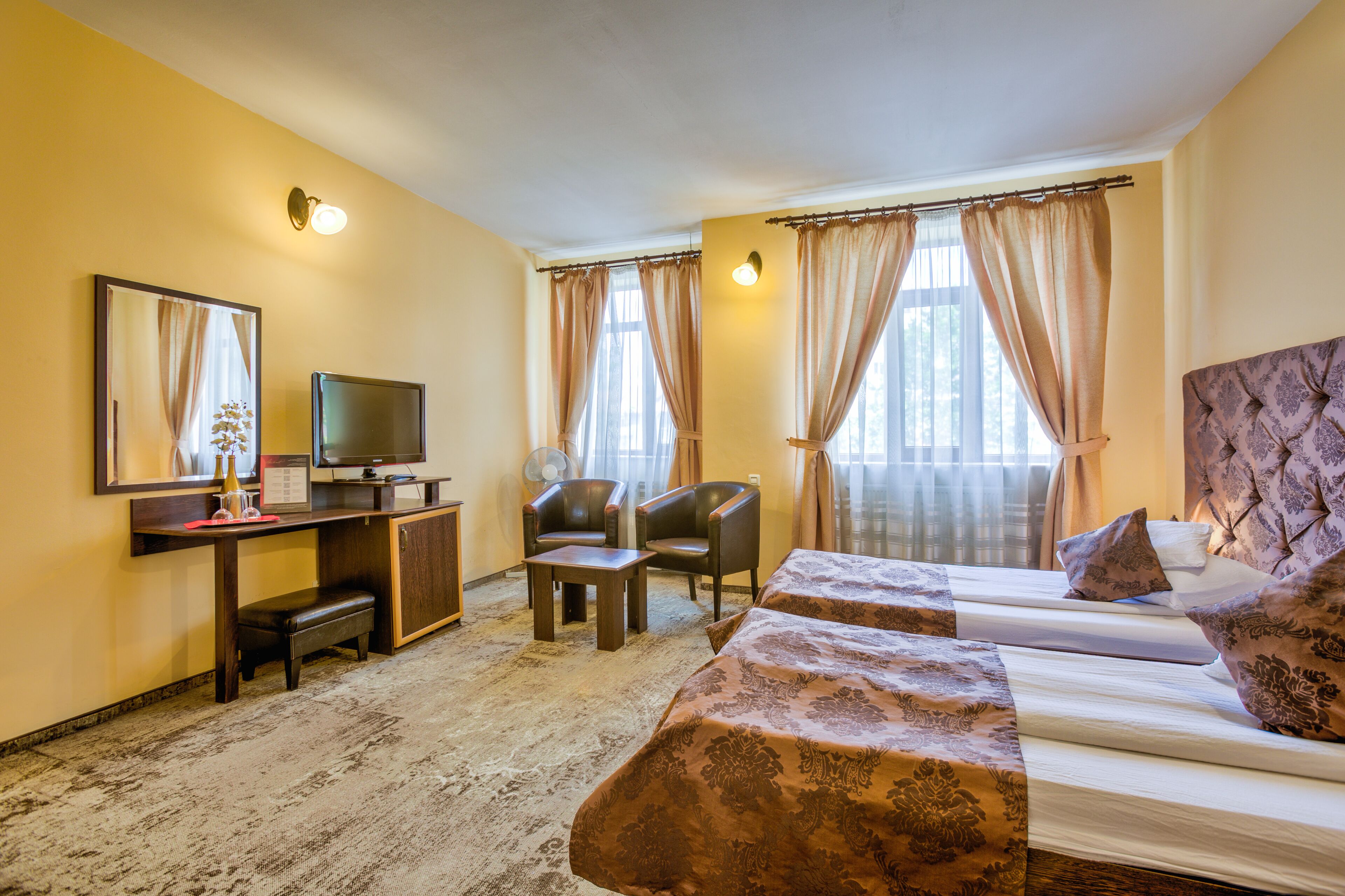 Quarto de hotel com cama, duas poltronas, escrivaninha com televisão e uma janela ampla com cortinas.