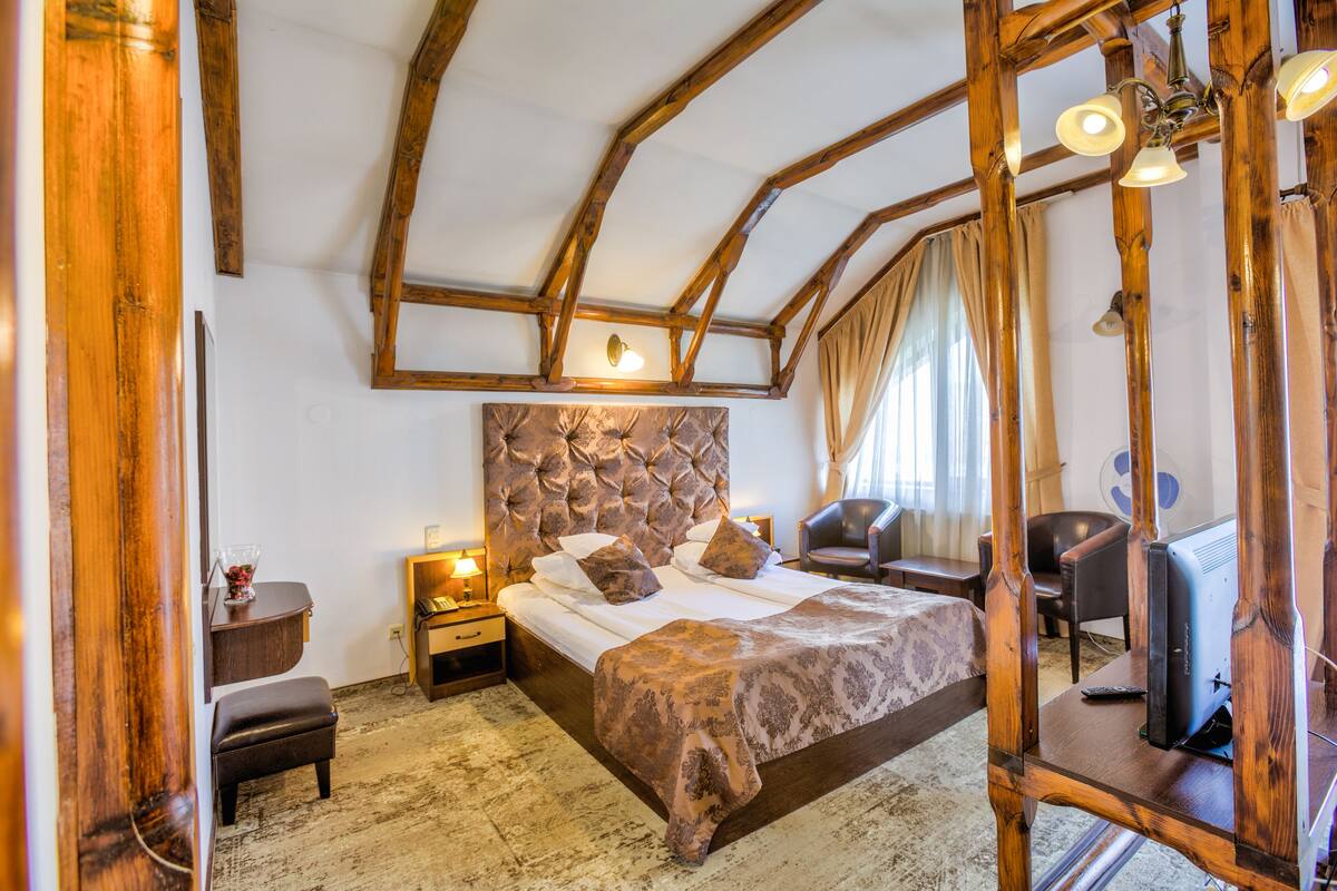 Un dormitorio con cabecera de madera, cama con ropa de cama estampada, mesita de noche con lámpara, una silla y un escritorio con computadora.