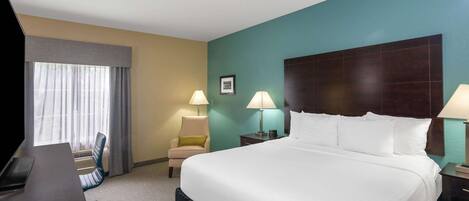 A hotel room with a large bed, a desk, a chair, two lamps, and a window with curtains.