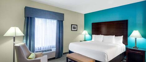 Una habitación de hotel con una cama grande, dos lámparas de noche, una silla, un banco y una ventana con cortinas.