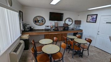 Una cafetería pequeña y limpia con mostrador, varias mesas y sillas. Se observan máquinas de café, un televisor y una puerta con la indicación 'Solo Personal'.
