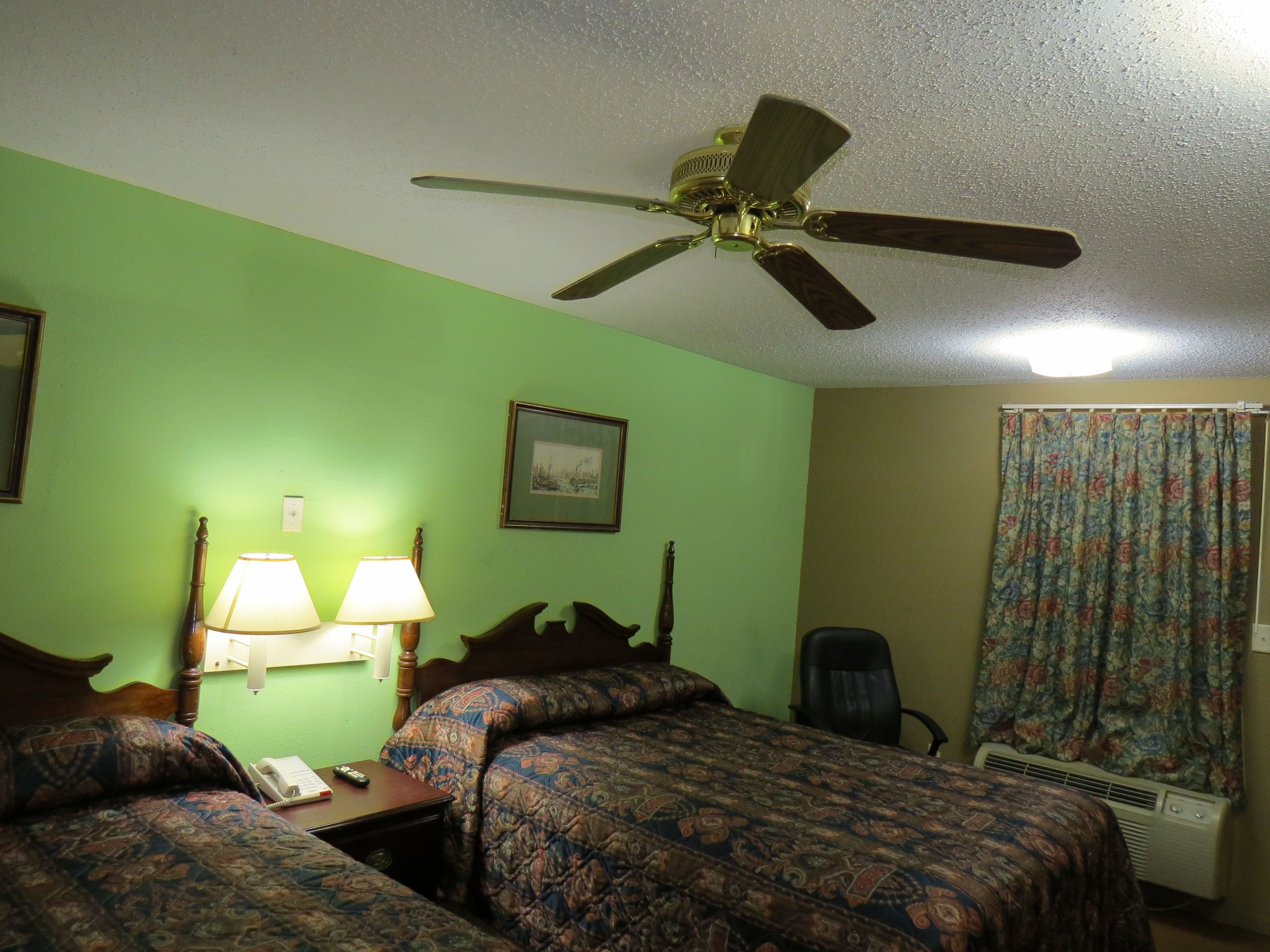 A hotel room with two beds, a ceiling fan, a desk with a lamp, a chair, and a window with curtains.