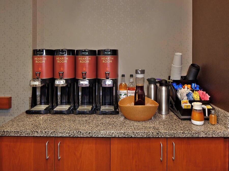 Una estación de café con cuatro dispensadores de marca, un cuenco con una botella, y diversos condimentos y utensilios.